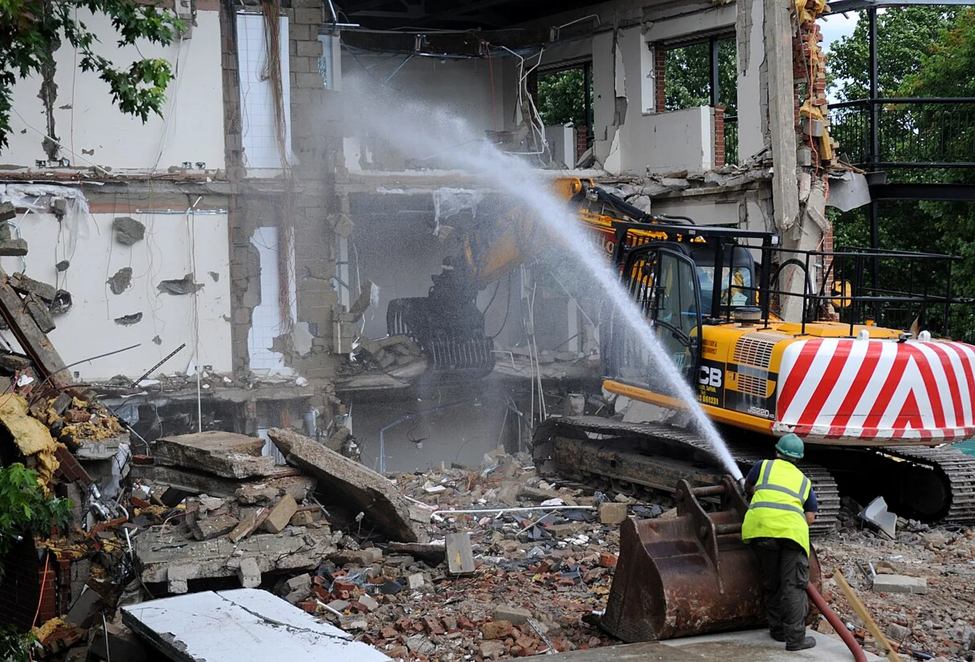 Demolition excavator working while a ground worker applies water for dust suppression outside the machine work area