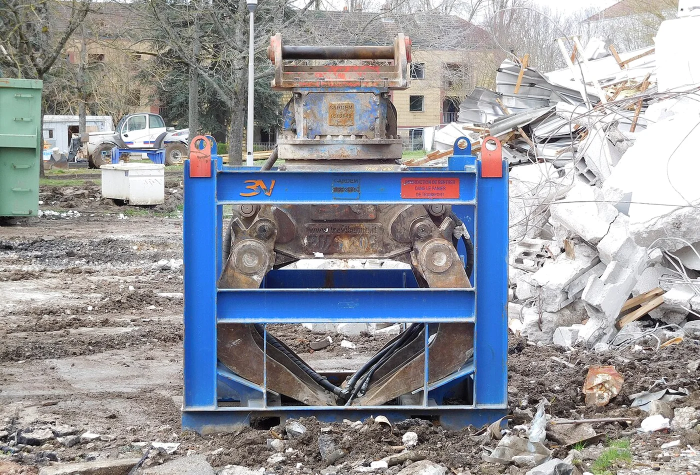 Demolition grapple secured upright in a steel transport and storage frame beside rubble